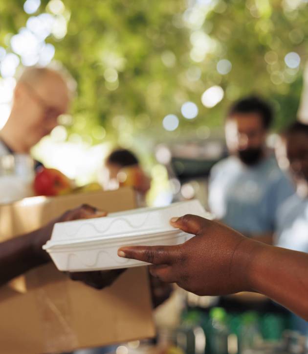 At food drive, volunteer with african american ethnicity serves a poor, hungry person a warm meal. Close-up of a less privileged needy individual receiving free food from charity worker.